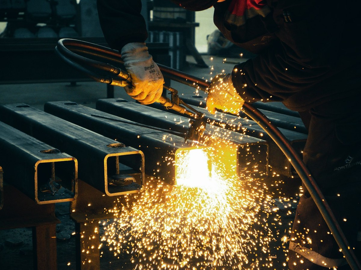 Bright fan of orange sparks flying as a welder cuts and shapes lengths of square steel tube on a rack