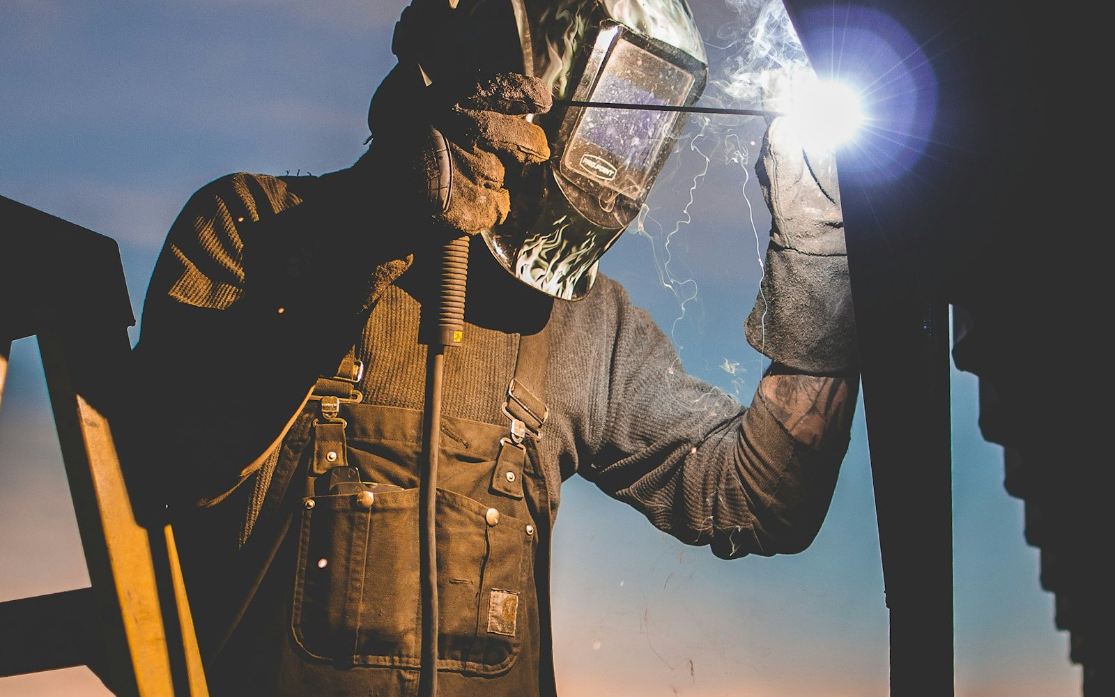 Welder in a helmet and work overalls striking a bright arc on steel at an outdoor jobsite against a sunset sky